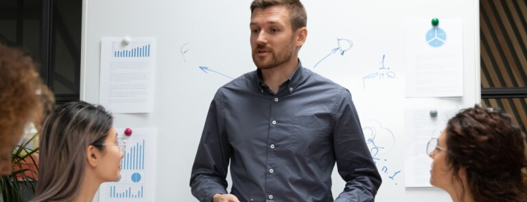 Man leads a team meeting standing in front of a white boar