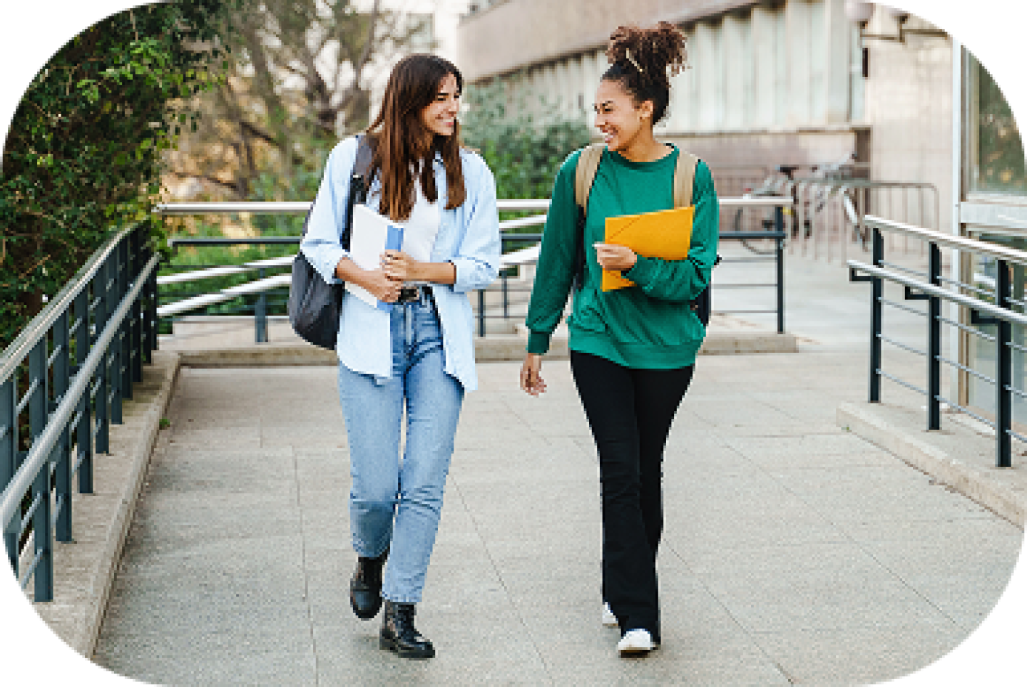 Two college students walking outdoors on campus, smiling and carrying notebooks and backpacks between classes.