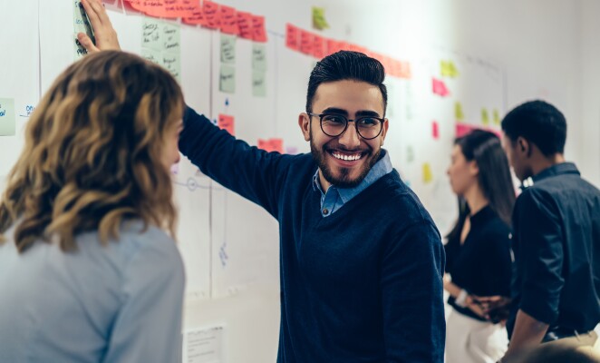 Man with a beard and glasses smiles at coworkers while working at the whiteboard