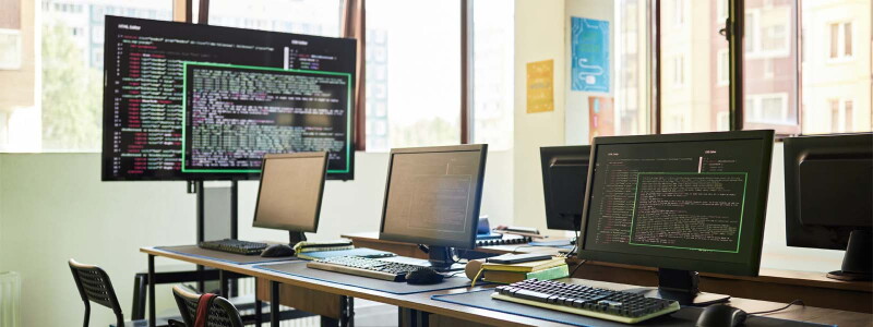 A sunny classroom is filled with various computers and screens displaying code.