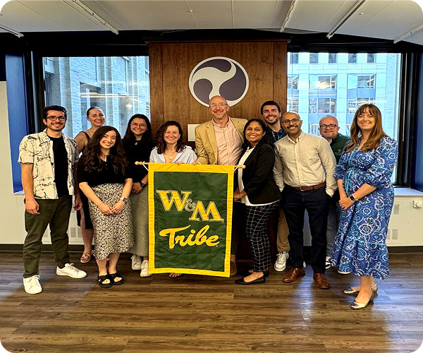 Group of people smiling indoors, holding a green and yellow "W&M Tribe" banner in front of a wooden wall with a circular logo.