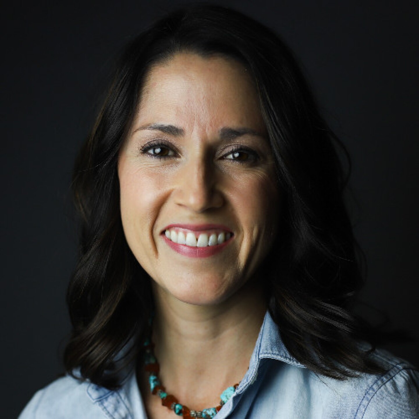 Professional headshot of a smiling woman with dark hair, wearing a light denim shirt and turquoise necklace against a dark background.