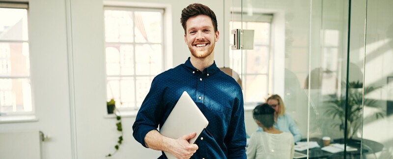 Smiling young businessman standing with his laptop in an office