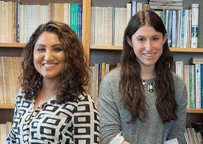 Two female students side by side in front of shelves of books