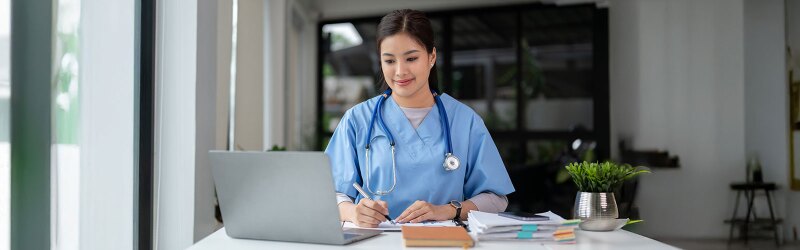 A nurse in scrubs is working on a laptop and taking notes.