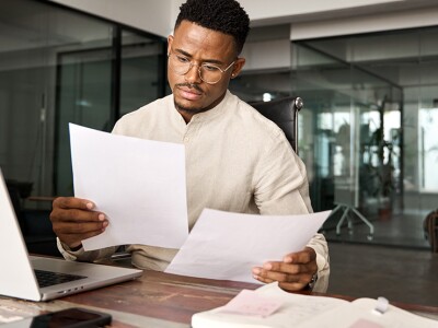 Man sits at a desk in front of an open laptop holding two sheets of paper and comparing information on the pages