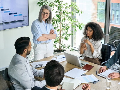 Three men and two women standing around a boardroom table with woman standing at the head of the table.
