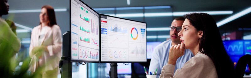 Professionals are gathered around two monitors displaying various business analytics charts and graphs, with one woman thoughtfully observing the data.
