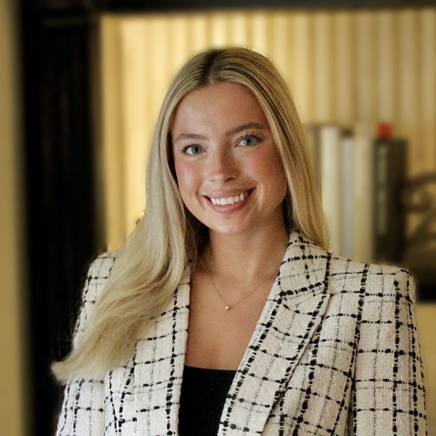 Professional headshot of Lexi Skirvin, with long blonde hair, wearing a white plaid blazer over a black top, against a softly blurred indoor background.