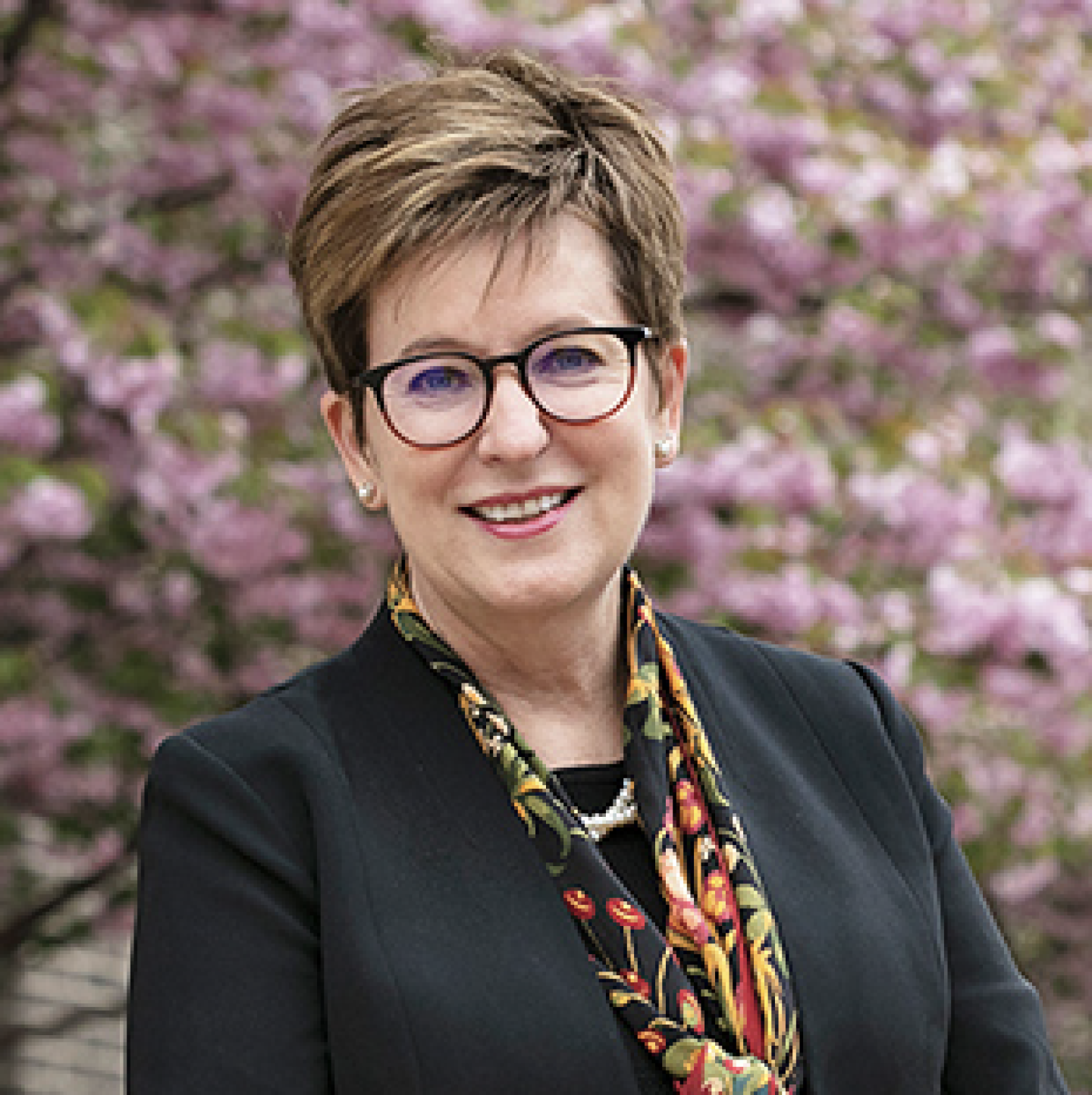 Professional headshot of a woman with short hair and glasses, wearing a dark blazer and floral scarf, smiling in front of blooming pink trees.