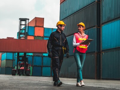 A man and a woman wearing hard hats walk through a work area filled with shipping crates.