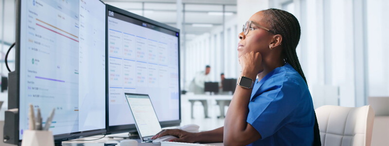 A woman in blue scrubs sits in front of a laptop and two external computer screens