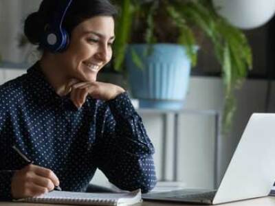 Smiling student wearing headphones and taking notes while seated at a laptop