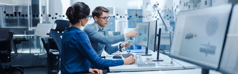 Two engineers collaborating in a modern lab, reviewing 3D mechanical designs on dual monitors while discussing a prototype part.