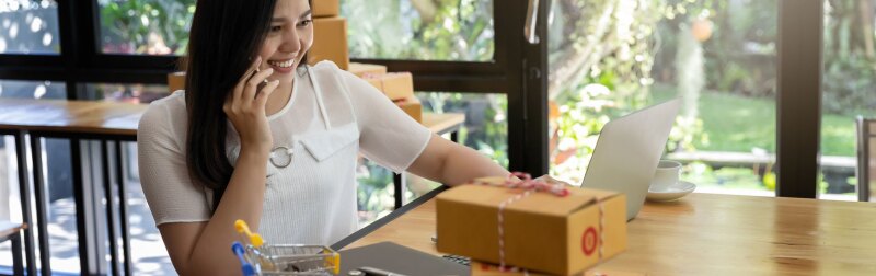 A seated, smiling woman talks on the phone while working on a laptop computer.