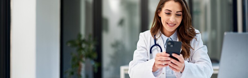 Female doctor sitting at the office table in front of the laptop and using a mobile phone