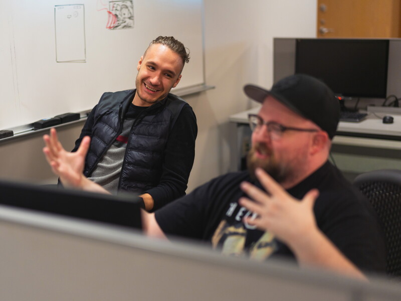 Two students engaged in conversation at a computer workstation in a classroom.