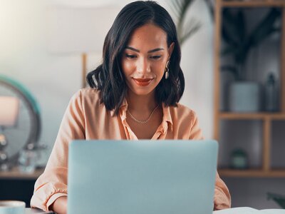 A smiling woman with dark hair sits at a desk, looking down at a laptop. A blurred room is in the background.