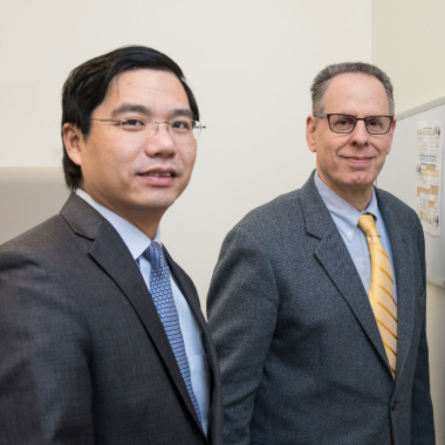Two male Yeshiva University professors smile while standing in an office with a whiteboard on the wall.