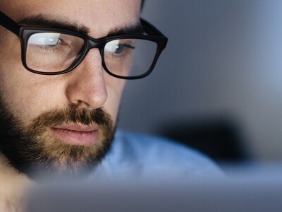 Man wearing glasses working with laptop in dark office late at night, his face lit up by screen