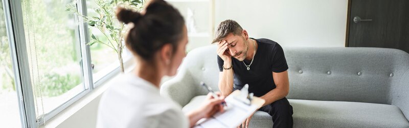 male client sitting on couch with head in hand while counselor takes notes