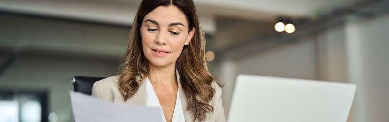 Business woman using a laptop in an office while looking over a paper letter