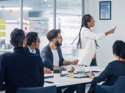 A diverse group of professionals in a marketing business meeting, a woman presents data on a large screen while colleagues observe and interact.