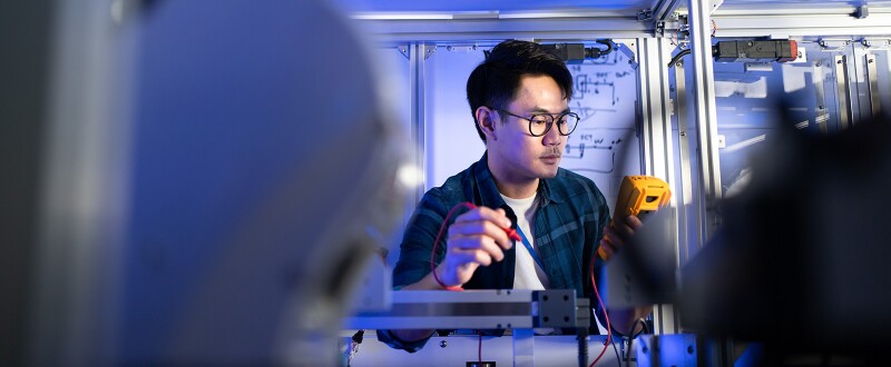 Electrical engineer using a digital multimeter to check voltage on an industrial machine, with a robotic machine in the foreground.