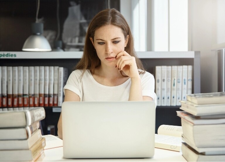 Woman in library with books around her, looking at a laptop
