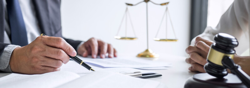 Hand holding fountain pen over business documents. Scales of justice is in the background, while a gavel is seen in the foreground.