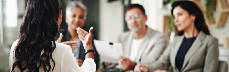 A woman with her back to the camera explains a concept to three other professionals.