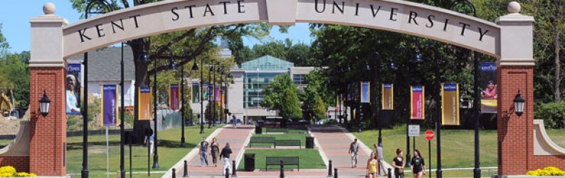 A view of a sunny day on the Kent State campus mall. A brick arch reads Kent State University. Students walk on brick paths among grassy grounds.