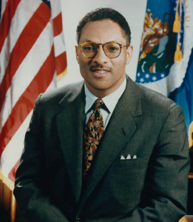 Man in suit leaning on desk in front of two flags
