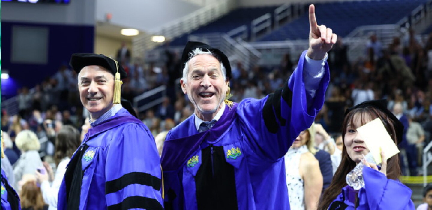 A male Hofstra Law graduate smiling with finger pointed in air walking with other graduates.