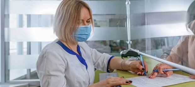 A masked woman sits on one side of a plexiglass divider reviewing a form with another person.