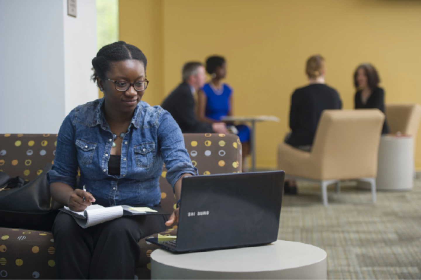 woman sitting on an office couch taking notes and looking at her laptop
