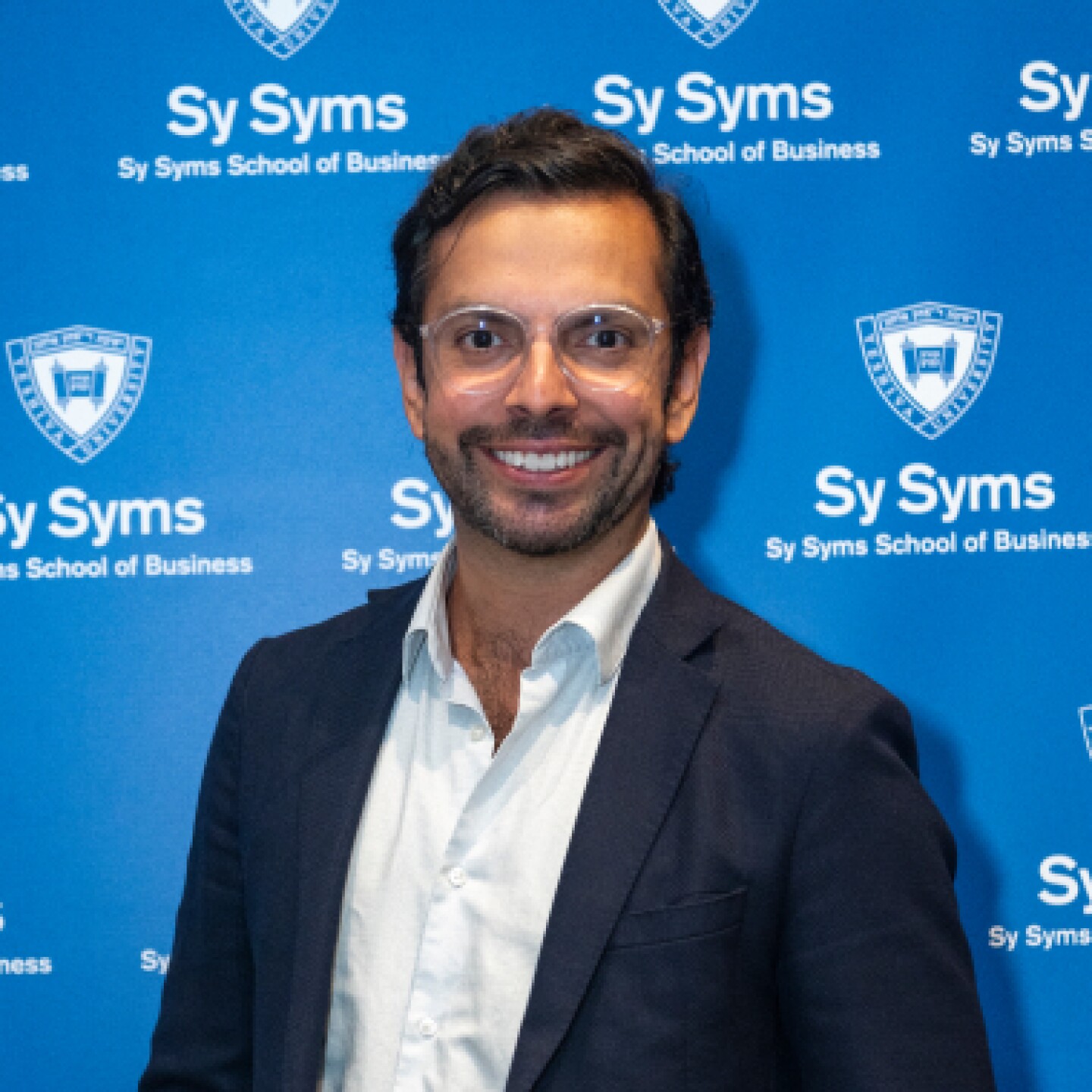 Smiling professional male stands in front of Yeshiva University Sy Syms backdrop.