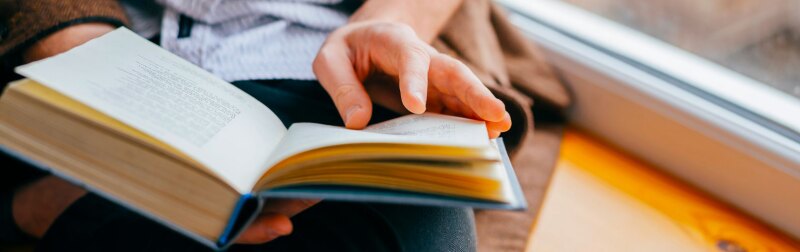Young man reading a book in his lap while sitting next to a window.