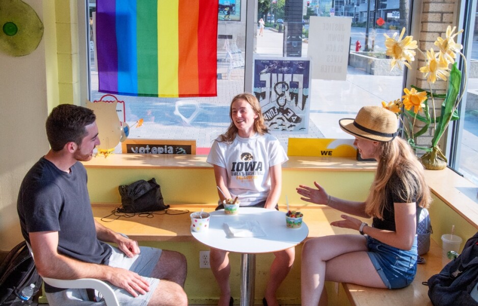 Three Iowa students seated and talking around table with pride flag on the window