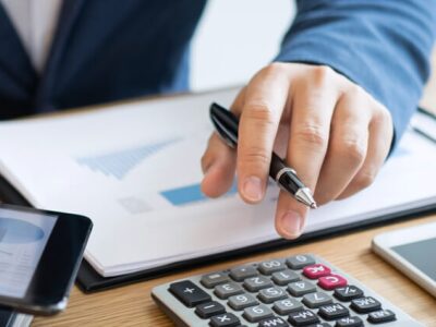 Hands of accountant in suit using tablet with charts, clipboard and calculator at desk