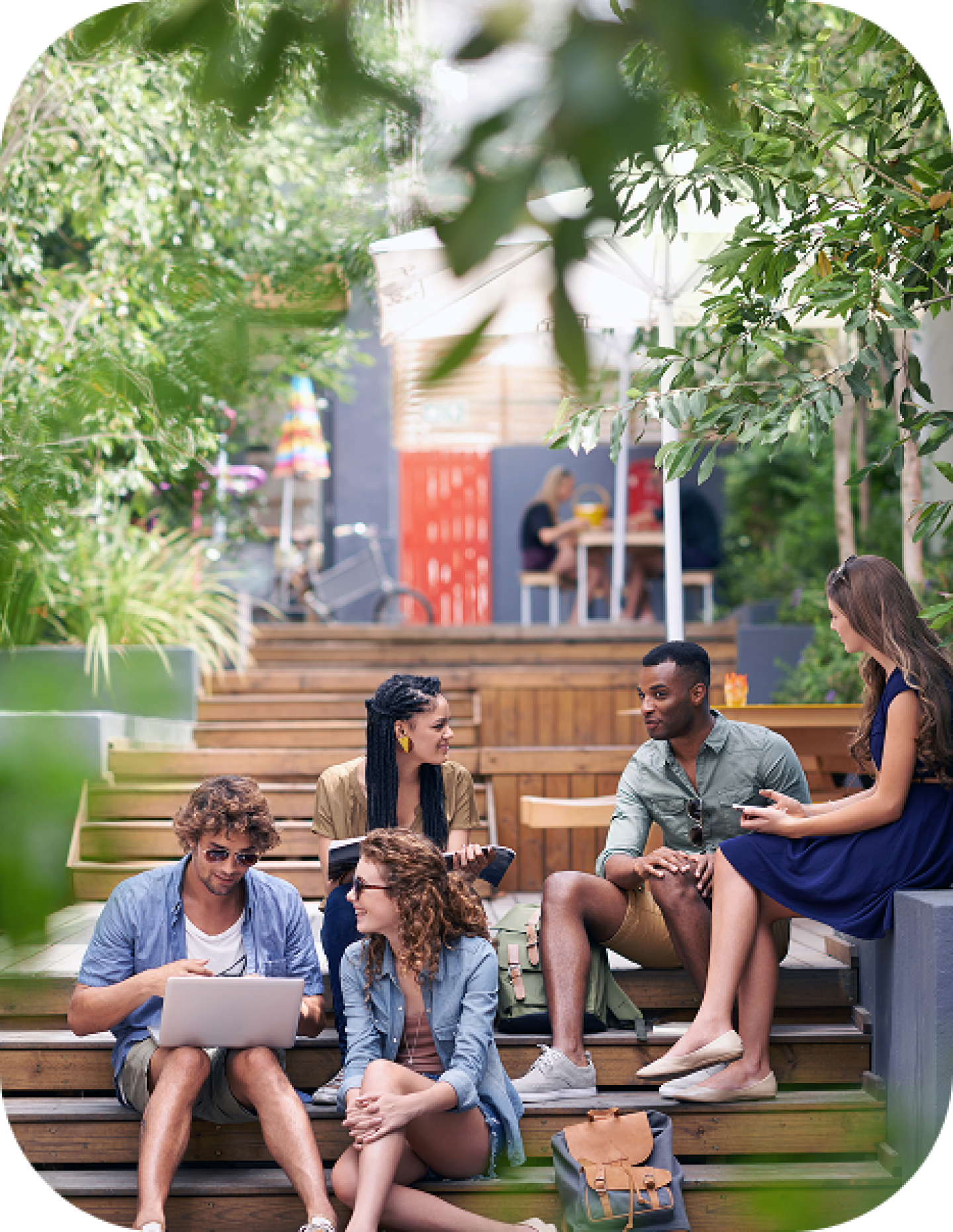 Group of college students sitting outdoors on campus steps, talking, studying, and using a laptop together.