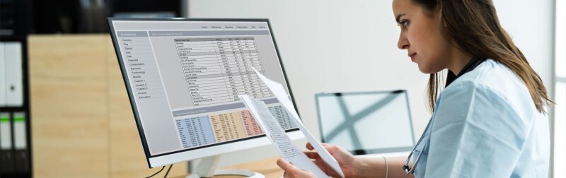 Female nursing manager sits at a desk in front of a computer as she reviews paper reports.