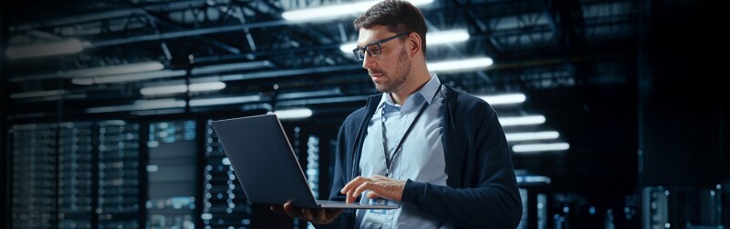 Man working on a laptop in a data center, surrounded by server racks
