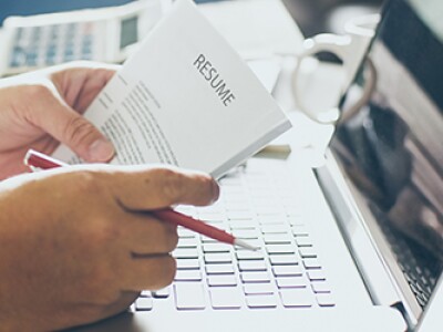 Hands holding a resume and a red pen while resting on a desk with a laptop