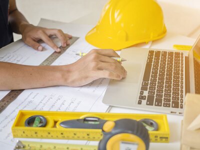 Civil engineer working with blueprints on a desk in front of a laptop surrounded by measuring tools and safety helmet