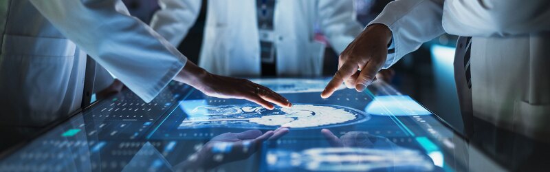 Doctors Interacting With Touch Screen On Table With MRI Scans Of Brain On Display.