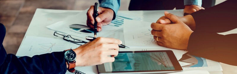 Hands of three people collaborating at a table over graphs and tablet
