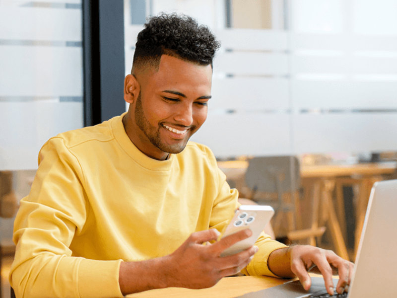 Smiling man in yellow sweatshirt looking at cellphone and working on laptop