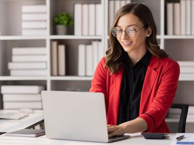smiling woman in red blazer sitting in white office space typing on laptop