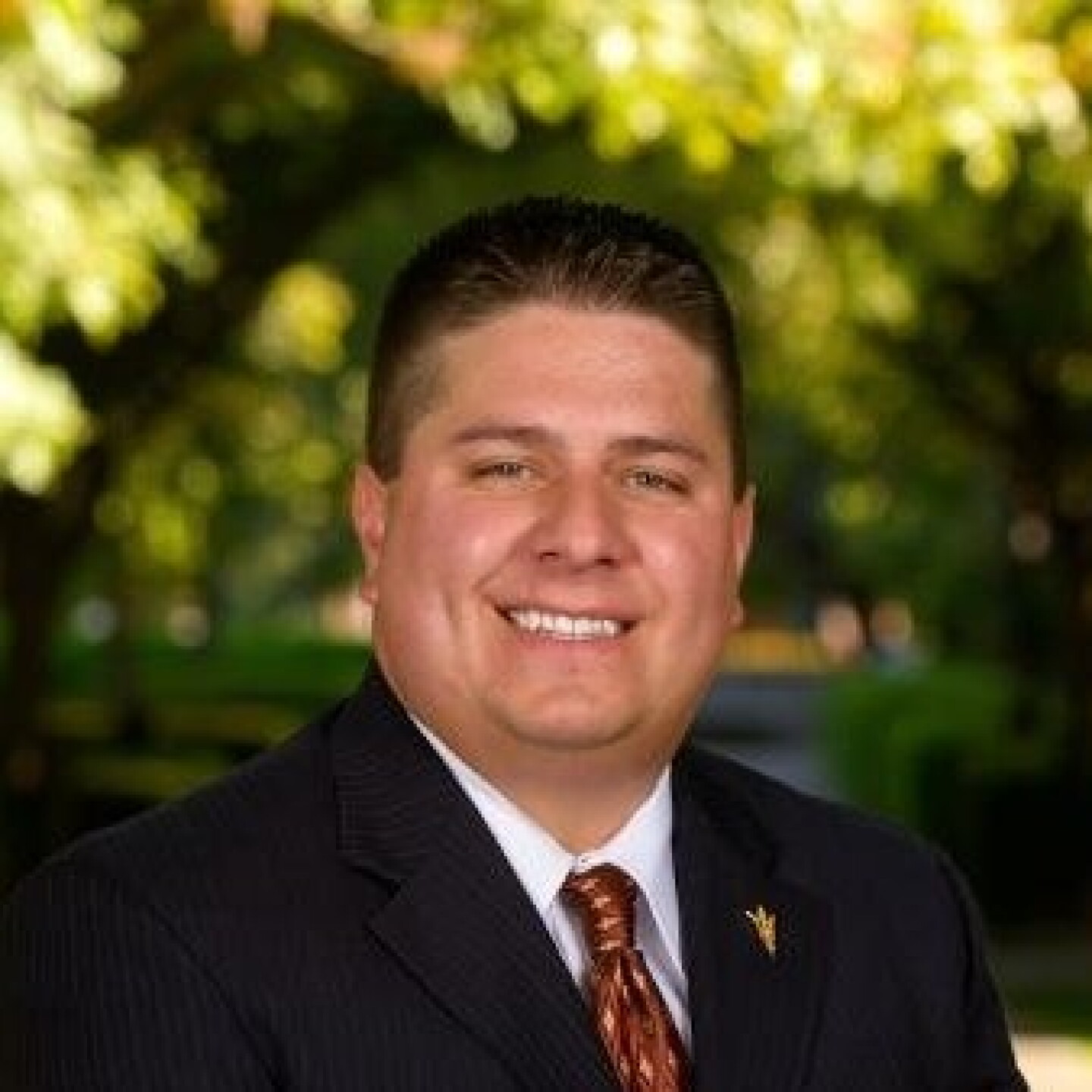 Smiling man in a dark suit and patterned tie outdoors, with sunlit green trees in the background.
